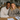 A diverse young couple, a Black man and a White woman, are seated together at a wooden table, smiling warmly at the viewer. The man on the left wears a cream cable knit sweater, and the woman on the right wears a brown knit sweater. On the table in front of them are several white documents, a pen, and autumn decorations including small orange and white pumpkins, and dried flowers. The background shows a cozy, light-filled home interior with shelves and plants.