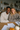 A diverse young couple, a Black man and a White woman, are seated together at a wooden table, smiling warmly at the viewer. The man on the left wears a cream cable knit sweater, and the woman on the right wears a brown knit sweater. On the table in front of them are several white documents, a pen, and autumn decorations including small orange and white pumpkins, and dried flowers. The background shows a cozy, light-filled home interior with shelves and plants.