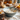 Close-up of a blue bowl filled with flour and a whisk resting inside. Around the bowl, there are cracked eggshells and scattered flour on the countertop. In the background, you can see whole eggs in a carton and small orange and yellow pumpkins along with autumn flowers, all set in a rustic kitchen.