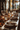 A long wooden table is set for a fall dinner, showcasing a festive and elegant atmosphere. Autumn leaves and miniature pumpkins are scattered amongst layered plates, woven placemats, and crystal glassware. Burning candles in ornate holders add a warm glow to the scene, creating a cozy and inviting ambiance.