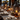 A long, wooden table is set for a fall dinner. Plates with folded linen napkins and silverware are arranged on each placemat. Small pumpkins, autumn leaves, rustic lanterns containing lit candles, and various glasses decorate the center of the table. The background features a dark window with out-of-focus lights.