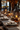 A long, wooden table is set for a fall dinner. Plates with folded linen napkins and silverware are arranged on each placemat. Small pumpkins, autumn leaves, rustic lanterns containing lit candles, and various glasses decorate the center of the table. The background features a dark window with out-of-focus lights.
