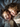 Close-up top-down shot of two smiling children lying on a gray bedspread. The child on the left has short, light brown hair and wears a patterned onesie. The child on the right has longer, curly brown hair and also wears a patterned onesie. Sunlight highlights their faces, revealing bright smiles and a warm, intimate moment.