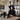 A young woman dressed as an elegant witch stands in a modern kitchen adorned with Halloween decorations. She has long dark hair, wears a black velvet dress with lace details, a classic black pointy witch hat, and ornate dark jewelry, looking directly at the viewer with a thoughtful expression. The kitchen features light marble-look counters with decorative human skulls, a lit candle in a dark holder, and a large black pot on the stove, creating a mysterious and sophisticated festive scene.