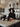 A young woman dressed as an elegant witch stands in a modern kitchen adorned with Halloween decorations. She has long dark hair, wears a black velvet dress with lace details, a classic black pointy witch hat, and ornate dark jewelry, looking directly at the viewer with a thoughtful expression. The kitchen features light marble-look counters with decorative human skulls, a lit candle in a dark holder, and a large black pot on the stove, creating a mysterious and sophisticated festive scene.