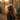 A person with long, voluminous, golden-brown curly hair is viewed from behind, wearing a dark brown coat. They are standing outdoors in an autumnal setting, with numerous orange and yellow leaves falling around them and scattered on the ground. Golden sunlight filters through the trees in the softly blurred background, creating a warm glow and lens flare.