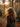 A person with long, voluminous, golden-brown curly hair is viewed from behind, wearing a dark brown coat. They are standing outdoors in an autumnal setting, with numerous orange and yellow leaves falling around them and scattered on the ground. Golden sunlight filters through the trees in the softly blurred background, creating a warm glow and lens flare.