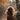 A vertical image showing the back of a person with voluminous, curly brown hair, glowing with golden backlight. They wear a dark brown coat and walk on a path covered in fallen autumn leaves, with more leaves suspended in the air. The background features a soft bokeh of sunlit trees and foliage in warm autumnal tones.