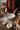 A close-up shot of a person wearing a rust-colored knit sweater, with their hands holding a whisk in a large cream-colored bowl containing white flour and a yellow liquid. The scene is set on a wooden table, decorated with small white pumpkins, dried brown leaves, and a cream lace placemat, evoking a warm and autumnal baking atmosphere.