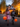 A vertical shot of two smiling children, a girl in a pink spiderweb dress and a boy in a skeleton costume, running down a paved street at dusk. They are holding orange pumpkin candy buckets, with a background of glowing warm string lights on autumn trees and a house, creating a magical Halloween scene.