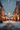 A vertical photo depicting a snow-covered city street at night, beautifully lit by numerous warm, golden Christmas lights. Softly falling snow creates a bokeh effect throughout the scene, contrasting with the cool blue-gray tones of the sky and the white snow on the ground. Buildings and decorated trees line both sides of the street, blurred into the background, adding to the overall atmospheric and enchanting winter scene.