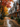 A picturesque autumn street scene features a row of modern townhouses on the right, with varying dark grey, brown, and reddish-brown siding and stone foundations. Trees displaying vibrant red, orange, and yellow foliage line the left side, and a multitude of fallen leaves are scattered across a wet, reflective pathway that leads into the distance. The overall mood is tranquil and serene under an overcast sky.