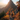 Two hikers, seen from behind, walk along a narrow, rocky mountain path that curves slightly. The path is surrounded by dense autumn foliage in brilliant shades of red, orange, and yellow, with some evergreen trees visible. In the background, a dramatic, jagged mountain peak is sharply silhouetted against a bright, warm sun low on the horizon, casting a glowing light over the scene and highlighting misty distant mountain ranges.