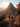 Two hikers, seen from behind, walk along a narrow, rocky mountain path that curves slightly. The path is surrounded by dense autumn foliage in brilliant shades of red, orange, and yellow, with some evergreen trees visible. In the background, a dramatic, jagged mountain peak is sharply silhouetted against a bright, warm sun low on the horizon, casting a glowing light over the scene and highlighting misty distant mountain ranges.