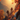 Two figures, seen from behind with backpacks, are hiking up a steep, rocky mountain path. The scene is illuminated by a brilliant golden-orange sunset, casting a warm glow over the landscape. Vibrant red and orange autumn foliage is prominent on trees and grasses along the path, with blurred leaves in the foreground, and distant mountains visible through a warm haze.