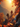 Two figures, seen from behind with backpacks, are hiking up a steep, rocky mountain path. The scene is illuminated by a brilliant golden-orange sunset, casting a warm glow over the landscape. Vibrant red and orange autumn foliage is prominent on trees and grasses along the path, with blurred leaves in the foreground, and distant mountains visible through a warm haze.