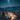 A breathtaking high-angle view of a snow-covered village at night, nestled within a valley surrounded by majestic, snow-capped mountains. The village is adorned with numerous warm, glowing Christmas lights on houses and evergreen trees, illuminating the pristine white landscape below a vibrant starry sky where the Milky Way is clearly visible. The scene evokes a magical, serene winter wonderland.