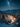 A breathtaking high-angle view of a snow-covered village at night, nestled within a valley surrounded by majestic, snow-capped mountains. The village is adorned with numerous warm, glowing Christmas lights on houses and evergreen trees, illuminating the pristine white landscape below a vibrant starry sky where the Milky Way is clearly visible. The scene evokes a magical, serene winter wonderland.
