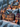 A close-up vertical shot of a beautifully detailed miniature Christmas village, covered in soft white snow. The ceramic buildings are lit from within, casting warm yellow and orange light from their many windows, and feature intricate designs, tiny figures, and miniature streetlights. Evergreen trees dot the snowy landscape, and the background is softly blurred with bokeh lights, creating a magical holiday ambiance.