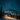 A vertical image depicting a cozy, illuminated wooden log cabin situated in a snow-covered mountain landscape at night. The cabin's warm, golden light spills onto the surrounding fresh snow and the trunks of nearby evergreen trees, all beneath a vast, dark blue sky teeming with countless sparkling stars. A large, snow-capped mountain rises majestically in the background, completing this serene and magical winter scene.