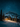 A vertical image depicting a cozy, illuminated wooden log cabin situated in a snow-covered mountain landscape at night. The cabin's warm, golden light spills onto the surrounding fresh snow and the trunks of nearby evergreen trees, all beneath a vast, dark blue sky teeming with countless sparkling stars. A large, snow-capped mountain rises majestically in the background, completing this serene and magical winter scene.