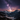 A breathtaking vertical landscape photograph captures the Milky Way galaxy arcing across a dark night sky with vibrant purple and orange hues near the horizon. Below, a vast valley is framed by towering snow-covered mountain peaks and dense, dark evergreen forests, leading down to a small, brightly lit village nestled in the snow. The scene evokes a sense of serene winter wilderness under a majestic starry sky, highlighting the beauty of nature and the cosmos.