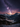 A breathtaking vertical landscape photograph captures the Milky Way galaxy arcing across a dark night sky with vibrant purple and orange hues near the horizon. Below, a vast valley is framed by towering snow-covered mountain peaks and dense, dark evergreen forests, leading down to a small, brightly lit village nestled in the snow. The scene evokes a sense of serene winter wilderness under a majestic starry sky, highlighting the beauty of nature and the cosmos.