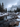 A vertical photograph capturing a serene winter mountain landscape. A clear river, speckled with visible rocks, winds through the snow-covered valley in the foreground and midground, flanked by tall evergreen pine trees. In the background, majestic snow-capped mountains rise into an overcast, cloudy sky, with some peaks partially obscured by mist, creating a peaceful and rugged scene.