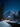 A breathtaking vertical shot of a winter night landscape. A deep blue, star-filled sky stretches above snow-capped mountains and a winding river in the foreground. Several small, warmly lit cabins are nestled among snow-dusted evergreen trees, creating a cozy focal point in the vast, snowy wilderness.