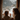 A vertical, natural light photograph showing a mother and young child from behind, looking out a rustic wooden window at a snow-covered mountain landscape. The child, with curly light brown hair, and the adult, with long wavy brown hair, both wear chunky knit grey sweaters and have their hands gently pressed against the cold window glass, sharing a moment of quiet wonder and connection in a warm, cozy interior setting.