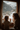 A vertical, natural light photograph showing a mother and young child from behind, looking out a rustic wooden window at a snow-covered mountain landscape. The child, with curly light brown hair, and the adult, with long wavy brown hair, both wear chunky knit grey sweaters and have their hands gently pressed against the cold window glass, sharing a moment of quiet wonder and connection in a warm, cozy interior setting.