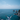 A person stands on a rocky cliff overlooking a vast blue ocean under a clear sky. The person, seen from behind, wears a white long-sleeved shirt, khaki shorts, and a blue baseball cap, with arms outstretched towards the sea. Jagged grey rock formations and small islands emerge from the deep blue water, conveying a powerful sense of freedom and awe amidst a stunning coastal landscape.