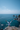 A person stands on a rocky cliff overlooking a vast blue ocean under a clear sky. The person, seen from behind, wears a white long-sleeved shirt, khaki shorts, and a blue baseball cap, with arms outstretched towards the sea. Jagged grey rock formations and small islands emerge from the deep blue water, conveying a powerful sense of freedom and awe amidst a stunning coastal landscape.