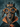 A close-up, eye-level portrait of a black and tan dachshund dog dressed in a whimsical Halloween costume. The dog wears large, round orange-framed glasses, a black glitter crown with the word 'BOO' in orange letters, and a black cape with an orange collar and bow. The background is a blurred, textured dark blue.