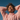 A young woman of color with curly dark hair and warm skin tones smiles joyfully, eyes closed, as golden sunlight illuminates her face. She wears a chunky light pink knit sweater and has her hands casually clasped behind her head, looking upwards against a clear, deep blue sky. The image conveys a feeling of peace, freedom, and pure happiness.