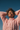 A young woman of color with curly dark hair and warm skin tones smiles joyfully, eyes closed, as golden sunlight illuminates her face. She wears a chunky light pink knit sweater and has her hands casually clasped behind her head, looking upwards against a clear, deep blue sky. The image conveys a feeling of peace, freedom, and pure happiness.