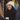 A young woman with dark hair, wearing a white knit beanie, a black coat, and black leather gloves, smiles brightly while holding a white coffee cup. She is seen in a medium close-up, engaged in conversation, against a blurred background of warm, golden string lights, suggesting a festive outdoor or urban winter setting. The image conveys a cheerful and cozy mood, with hints of holiday decorations.
