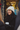A young woman with dark hair, wearing a white knit beanie, a black coat, and black leather gloves, smiles brightly while holding a white coffee cup. She is seen in a medium close-up, engaged in conversation, against a blurred background of warm, golden string lights, suggesting a festive outdoor or urban winter setting. The image conveys a cheerful and cozy mood, with hints of holiday decorations.