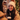 A young woman with dark wavy hair, wearing a cream knit beanie and a beige puffer jacket, smiles broadly and genuinely while holding a light brown disposable coffee cup in her black-gloved hands. She looks towards another person, whose blurred shoulder is in the foreground, suggesting a friendly conversation. The background is softly blurred with warm, glowing Christmas lights and green garland, indicating a festive outdoor winter evening.