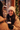 A young woman with dark wavy hair, wearing a cream knit beanie and a beige puffer jacket, smiles broadly and genuinely while holding a light brown disposable coffee cup in her black-gloved hands. She looks towards another person, whose blurred shoulder is in the foreground, suggesting a friendly conversation. The background is softly blurred with warm, glowing Christmas lights and green garland, indicating a festive outdoor winter evening.