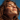A close-up, low-angle portrait shows a young woman with reddish-brown, windswept hair, her eyes gently closed and head tilted upwards, smiling softly. Her face is covered in natural freckles, illuminated by warm sunlight against a clear, vibrant blue sky. She appears to be enjoying a moment of peace and warmth outdoors.