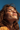 A close-up, low-angle portrait shows a young woman with reddish-brown, windswept hair, her eyes gently closed and head tilted upwards, smiling softly. Her face is covered in natural freckles, illuminated by warm sunlight against a clear, vibrant blue sky. She appears to be enjoying a moment of peace and warmth outdoors.