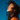A close-up, profile shot of a young woman with dark, wind-swept hair looking upwards with her eyes closed against a vibrant blue sky. Her skin is illuminated by warm sunlight, appearing dewy and radiant, and she wears a bright orange top. The image evokes a strong sense of peace and natural joy.