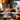 A warm-toned, eye-level photograph of a stainless steel pot on a stovetop, actively steaming with boiling water. To the right, on a wooden counter, are bundles of cinnamon sticks and a small orange decorative pumpkin, set on a rustic woven mat. The background is softly blurred, revealing autumn leaves in reds and oranges, hinting at a cozy kitchen scene.
