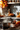 A warm-toned, eye-level photograph of a stainless steel pot on a stovetop, actively steaming with boiling water. To the right, on a wooden counter, are bundles of cinnamon sticks and a small orange decorative pumpkin, set on a rustic woven mat. The background is softly blurred, revealing autumn leaves in reds and oranges, hinting at a cozy kitchen scene.