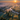 A detailed aerial photograph captures a breathtaking misty rural landscape at sunrise. The vibrant orange sun bursts from the upper right, casting long, dramatic shadows from numerous trees across fog-covered fields and a winding river below. The sky transitions from a deep blue at the top to a warm orange and yellow near the horizon, while golden crepuscular rays pierce through the thick mist, highlighting pockets of green and autumnal foliage around a small cluster of houses.