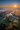 A detailed aerial photograph captures a breathtaking misty rural landscape at sunrise. The vibrant orange sun bursts from the upper right, casting long, dramatic shadows from numerous trees across fog-covered fields and a winding river below. The sky transitions from a deep blue at the top to a warm orange and yellow near the horizon, while golden crepuscular rays pierce through the thick mist, highlighting pockets of green and autumnal foliage around a small cluster of houses.