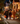 Three young children, dressed in Halloween costumes, walk down a dimly lit residential sidewalk at night. A boy in a skeleton costume holds an orange pumpkin bucket, a girl in a witch's dress smiles, and another child in a tiger costume carries a glowing lantern. The background features houses decorated with warm string lights and pumpkins, creating a festive, inviting scene.
