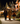 A photograph shows three young children in Halloween costumes walking side-by-side on a concrete sidewalk at night. From left, one child wears a black skeleton costume and holds a flashlight, the middle child wears a full orange and black tiger suit and holds an illuminated jack-o'-lantern bucket, and the child on the right wears a black and orange leopard print top with a tutu skirt and striped tights. In the blurred background, houses are adorned with warm, glowing Halloween decorations and pumpkins.