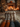 A low-angle shot depicts two pairs of legs, clad in intricately patterned knitted socks (one pair orange and grey, the other cream and brown), resting on a white faux fur rug. In the softly blurred background, a warm fire crackles in a rustic stone fireplace, flanked by stacked logs and illuminated by numerous flickering candles and warm string lights, creating an inviting and tranquil ambiance.